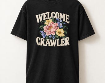 Può includere: Una t-shirt nera a maniche corte con una grafica centrale raffigurante un bouquet di fiori rosa, gialli e blu. Il testo WELCOME CRAWLER è stampato in un carattere serif arcuato color crema sopra e sotto la composizione floreale.