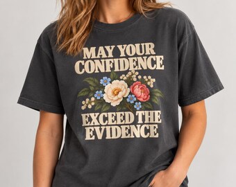 Può includere: Una t-shirt a maniche corte grigio antracite con il testo color crema "MAY YOUR CONFIDENCE EXCEED THE EVIDENCE" centrato sopra e sotto un'illustrazione colorata di fiori bianchi, rosa e blu con foglie verdi.