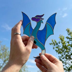 Könnte beinhalten: Ein Drachen-Sonnenfänger aus Buntglas mit einem violetten Kopf, einem türkisfarbenen Hals und Körper sowie Flügeln aus hell- und dunkelblauen Glassegmenten. Die einzelnen Glasteile sind durch dünne, dunkle Metalllötlinien miteinander verbunden.