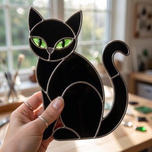 May include: A stained glass suncatcher shaped like a sitting cat with opaque black glass panels and vibrant translucent green glass eyes. The cat figure is outlined with silver-toned metal solder lines that define the ears, face, and curved tail.