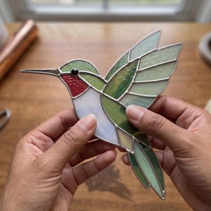 May include: A stained glass hummingbird suncatcher featuring a red throat, white chest, and green wings constructed from multiple glass segments joined by silver metal solder lines, with a small black bead serving as the bird's eye.