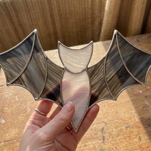 May include: A stained glass bat ornament featuring translucent white glass for the body and head, contrasted with dark, streaked grey glass for the wings. The individual glass segments are joined together with thin, silver-colored metal solder lines.