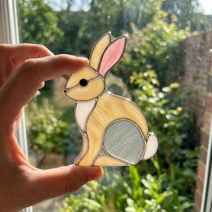 May include: A stained glass suncatcher shaped like a sitting rabbit. The rabbit features light tan body glass, a white chest, a pale pink inner ear, a textured grey hind leg, and a small white tail, all held together by dark metal solder lines.