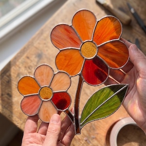 May include: A stained glass suncatcher featuring two flowers with orange, red, and yellow petals, a central yellow core, and a single green leaf. The glass pieces are joined by thin metal solder lines, creating a vibrant, translucent floral design.