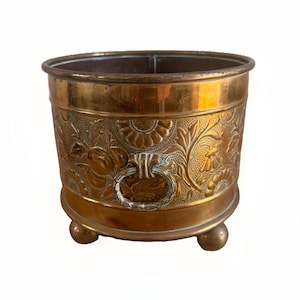 May include: A round brass planter featuring an embossed floral and fan pattern on the exterior. The vessel includes two side ring handles and rests on three small ball-shaped feet. The metal surface displays a warm, polished metallic finish.