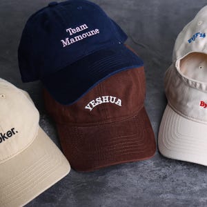 Op de afbeelding: Een collectie van vier baseballpetten in beige, marineblauw, bruin en off-white. De beige pet heeft de tekst 'Poker.', de marineblauwe pet zegt 'Team Mamoune', de bruine pet zegt 'YESHUA' en de off-white pet toont 'EVJF'24 - 20 to 22.09' en 'By Billie'.