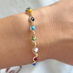 May include: A gold-toned wire bracelet featuring a series of small, multi-coloured evil eye beads. The beads are arranged in a vertical line, showcasing colours including yellow, black, light blue, green, white, red, and dark blue along the wire.