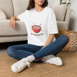 Könnte beinhalten: Ein weißes T-Shirt mit kurzen Ärmeln und einer Grafik einer roten Schüssel voller bunter Cerealien sowie einem silbernen Löffel. Unter der Schüssel steht der Text "Knusprig daneben" in einer bunten, abgerundeten Schrift, umgeben von Cerealien.
