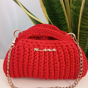 May include: A bright red crocheted handbag featuring a thick, ribbed texture and a gold-toned metal chain strap. The bag has a rectangular shape with rounded edges and a gold metal plaque on the front with the word HandMade written in script.