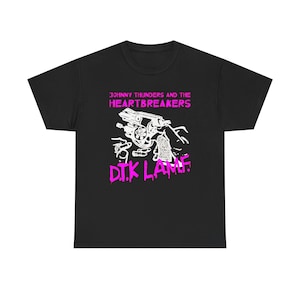 Peut inclure: Un t-shirt noir à manches courtes avec un motif blanc représentant une main tenant un revolver et des balles. Le texte JOHNNY THUNDERS AND THE HEARTBREAKERS et D.T.K L.A.M.F. est imprimé en rose vif au-dessus et en dessous de l'illustration.
