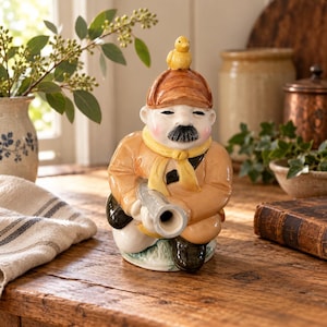 May include: A ceramic figurine of a person wearing a tan jacket, a yellow scarf, and a brown hat with a small yellow duck perched on top. The figure has a black moustache, rosy cheeks, and is holding a cylindrical object in a crouching position.