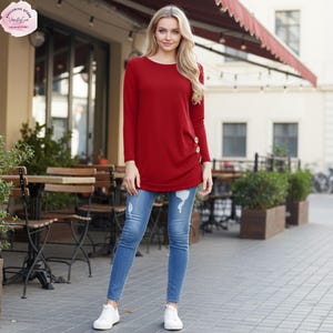 May include: A long-sleeved, deep red tunic top featuring a round neckline and a side-slit detail accented with three light-colored buttons. The top is paired with blue denim jeans that have distressed knee areas and white sneakers.