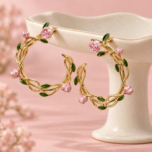 May include: A pair of gold hoop earrings featuring a vine-like design with small pink enamelled flowers and green enamelled leaves. The circular hoops are constructed from twisted metal wire, creating a delicate botanical appearance.