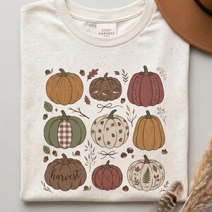 May include: A cream-colored t-shirt with a graphic print featuring nine stylized pumpkins in various autumn colors and patterns. The design includes small illustrations of leaves, acorns, and snails, with the word harvest written in cursive script.