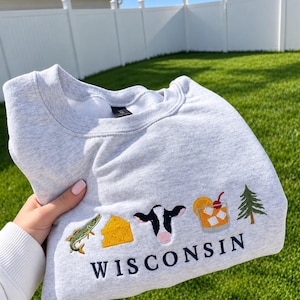 May include: A light gray heather crewneck sweatshirt featuring embroidered icons of a fish, a wedge of cheese, a cow head, an old fashioned cocktail with a cherry, and a pine tree, with the word WISCONSIN embroidered in dark blue below them.
