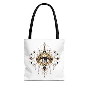 May include: A white tote bag with black handles featuring a central graphic of a stylized eye with a crescent moon pupil, surrounded by golden sun rays, celestial moon phases, and hanging beaded ornaments in a symmetrical arrangement.
