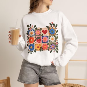 May include: A white long-sleeve sweatshirt featuring a central graphic of colorful crochet-style squares. The design includes various flowers, a red heart, and a strawberry, all surrounded by green leafy vines and small blossoms.