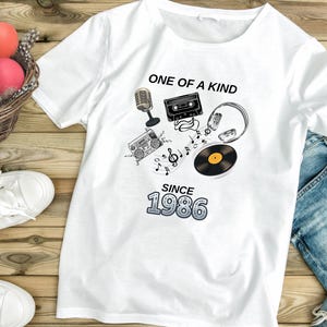 Könnte beinhalten: Ein weißes T-Shirt mit einem Grafikdruck aus Mikrofon, Kassette, Ghettoblaster, Kopfhörern und Schallplatte mit Noten. Der Text ONE OF A KIND SINCE 1986 ist in schwarzer und blauer Schrift mittig auf der Brust abgebildet.
