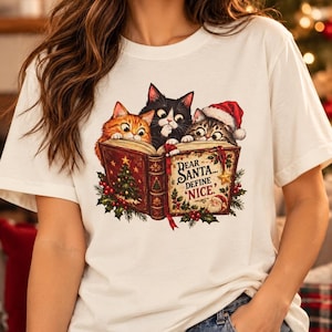 Op de afbeelding: Een wit t-shirt met een afbeelding van drie katten die in een open boek kijken. De boekomslag toont een kerstboom, hulst en de tekst: Dear Santa... Define 'Nice.' Eén kat draagt een rode kerstmuts met feestelijke details.