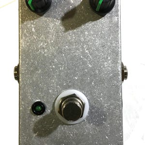 May include: A silver-colored guitar effects pedal with two black knobs featuring green indicator lines. It has a white footswitch, a green LED, and two input/output jacks. The pedal is rectangular and has a textured surface.