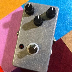 May include: A silver-colored effects pedal with three black knobs and a white footswitch. A small red light is visible near the footswitch. The pedal is rectangular and has a textured surface. The background is a colorful patchwork of felt.