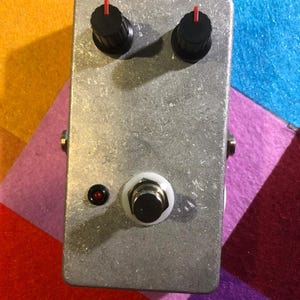 May include: A rectangular, silver-colored effects pedal with two black knobs with red indicators, a white footswitch, and a red LED indicator. The pedal is designed for use with electric guitars or other instruments.