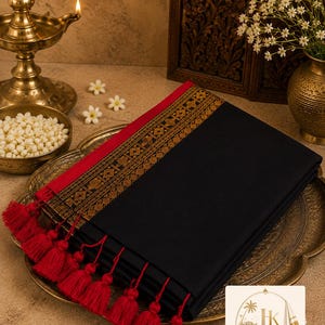 May include: A folded black textile featuring a wide red border with intricate gold geometric patterns and a row of red tassels along the edge. The fabric is neatly stacked on a metallic tray, showcasing a deep black colour and detailed embroidery.