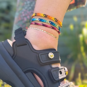 May include: Three layered anklets consisting of one thin gold chain and two woven fabric bands in orange, yellow, blue, and pink, worn above a black platform sandal featuring three adjustable straps with decorative flower-shaped metal charms.