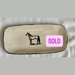 May include: A rectangular ceramic tray with a cream-colored glaze and black speckled details. The tray features a black line drawing of a horse in the center and a pink rectangular sticker with the word SOLD printed in white capital letters.