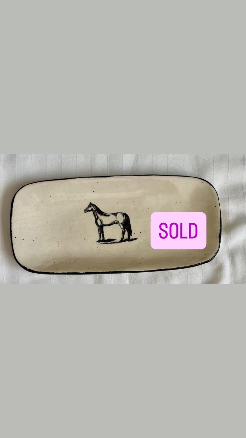 May include: A rectangular ceramic tray with a cream-colored glaze and black speckled details. The tray features a black line drawing of a horse in the center and a pink rectangular sticker with the word SOLD printed in white capital letters.
