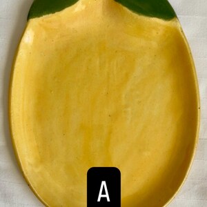 Könnte beinhalten: Eine zitronenförmige Keramikschale mit glänzend gelber Oberfläche und einem einfarbig grün bemalten oberen Bereich. Die Schale hat eine glatte, leicht konkave Form und ein spitz zulaufendes Ende. Ein schwarzes Etikett mit dem weißen Buchstaben A.
