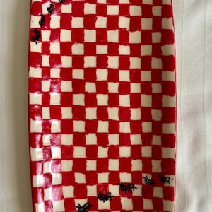 May include: A rectangular ceramic serving tray featuring a hand-painted red and white checkered pattern. The surface is decorated with small, black, hand-drawn ant illustrations that appear to crawl diagonally across the glossy, glazed finish.