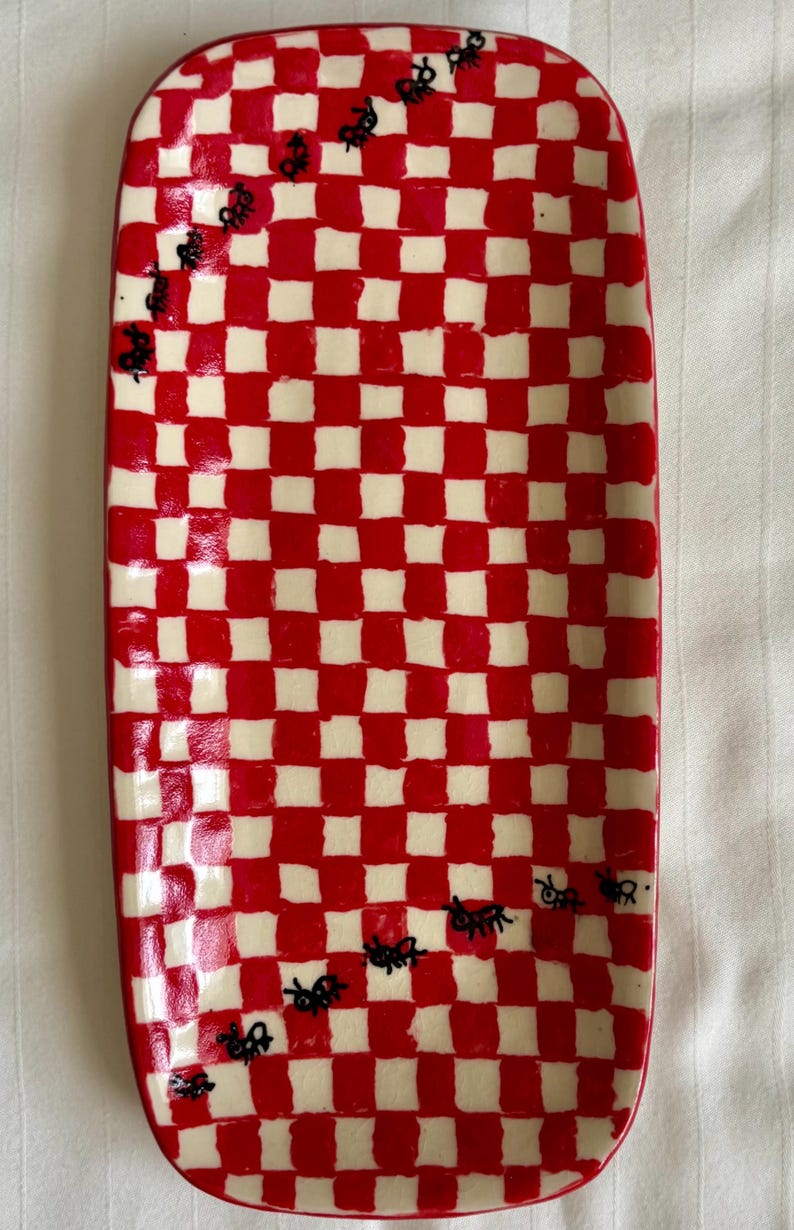 May include: A rectangular ceramic serving tray featuring a hand-painted red and white checkered pattern. The surface is decorated with small, black, hand-drawn ant illustrations that appear to crawl diagonally across the glossy, glazed finish.