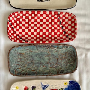 May include: Four rectangular ceramic trays arranged vertically. The top tray is cream with a black horse illustration. The second is a red and white checkerboard pattern. The third has a textured blue-green glaze. The bottom is cream with painted flowers, a butterfly, and a large red flower.