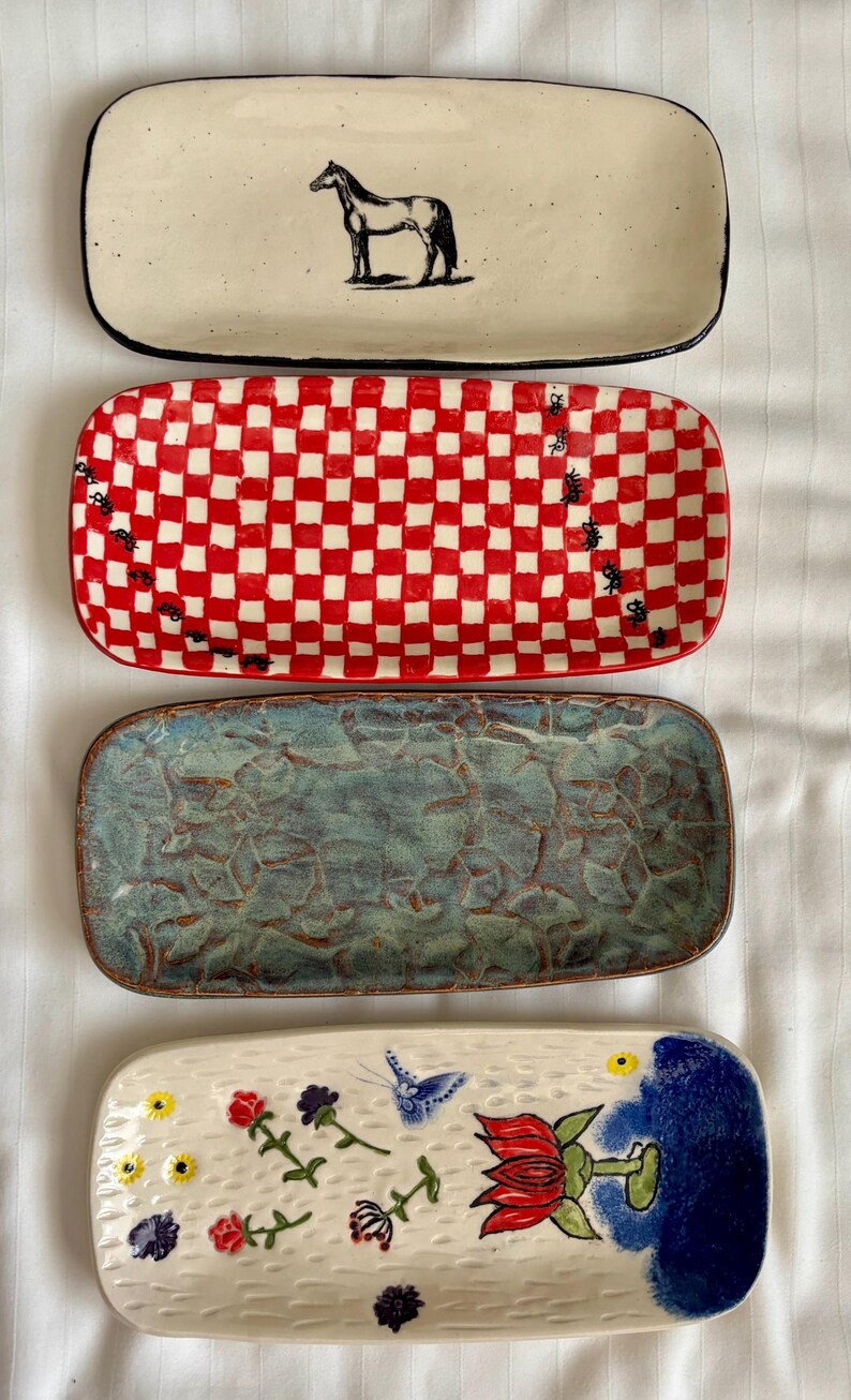 May include: Four rectangular ceramic trays arranged vertically. The top tray is cream with a black horse illustration. The second is a red and white checkerboard pattern. The third has a textured blue-green glaze. The bottom is cream with painted flowers, a butterfly, and a large red flower.