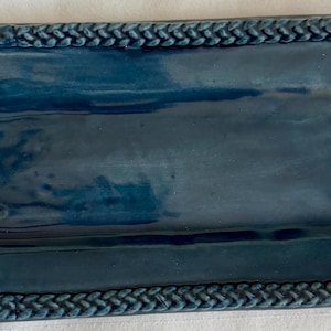 May include: A rectangular ceramic tray with a glossy deep blue finish. The tray features a raised border with a textured, braided rope pattern along all four edges, surrounding a smooth, flat, and slightly reflective central surface area.