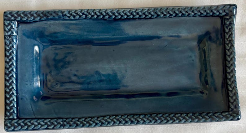 May include: A rectangular ceramic tray with a glossy deep blue finish. The tray features a raised border with a textured, braided rope pattern along all four edges, surrounding a smooth, flat, and slightly reflective central surface area.