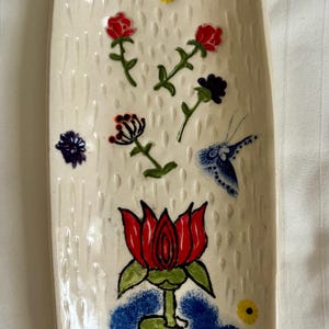 May include: A rectangular ceramic tray with a cream-colored base featuring a textured rain-drop pattern. The surface is decorated with hand-painted red, purple, and yellow flowers, a blue butterfly, and a large red lotus flower in blue water.