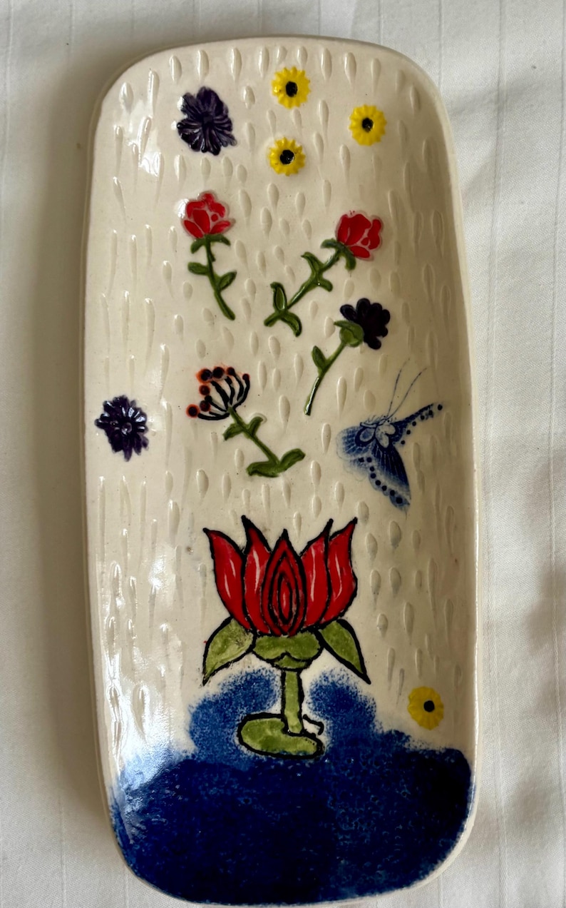 May include: A rectangular ceramic tray with a cream-colored base featuring a textured rain-drop pattern. The surface is decorated with hand-painted red, purple, and yellow flowers, a blue butterfly, and a large red lotus flower in blue water.