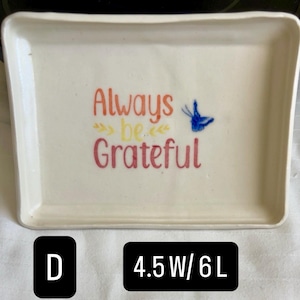 May include: A rectangular off-white ceramic tray featuring the orange and pink text Always be Grateful with yellow decorative arrows and a blue butterfly graphic. The tray measures 4.5 inches wide by 6 inches long as indicated by the text label D.