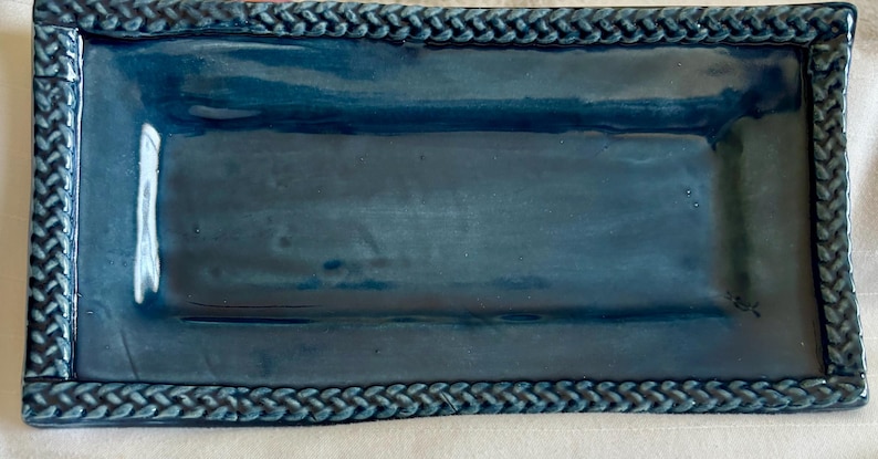 This is a handcrafted blue ceramic rectangular decorative tray featuring a textured braided rope border.  4 1/2 inch wide and 9 inch long.