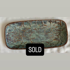 May include: A rectangular ceramic tray with rounded corners, featuring a mottled blue and green glaze with a textured, crackled surface pattern and a dark brown rim. A black rectangular label with the white text SOLD is centered on the tray.