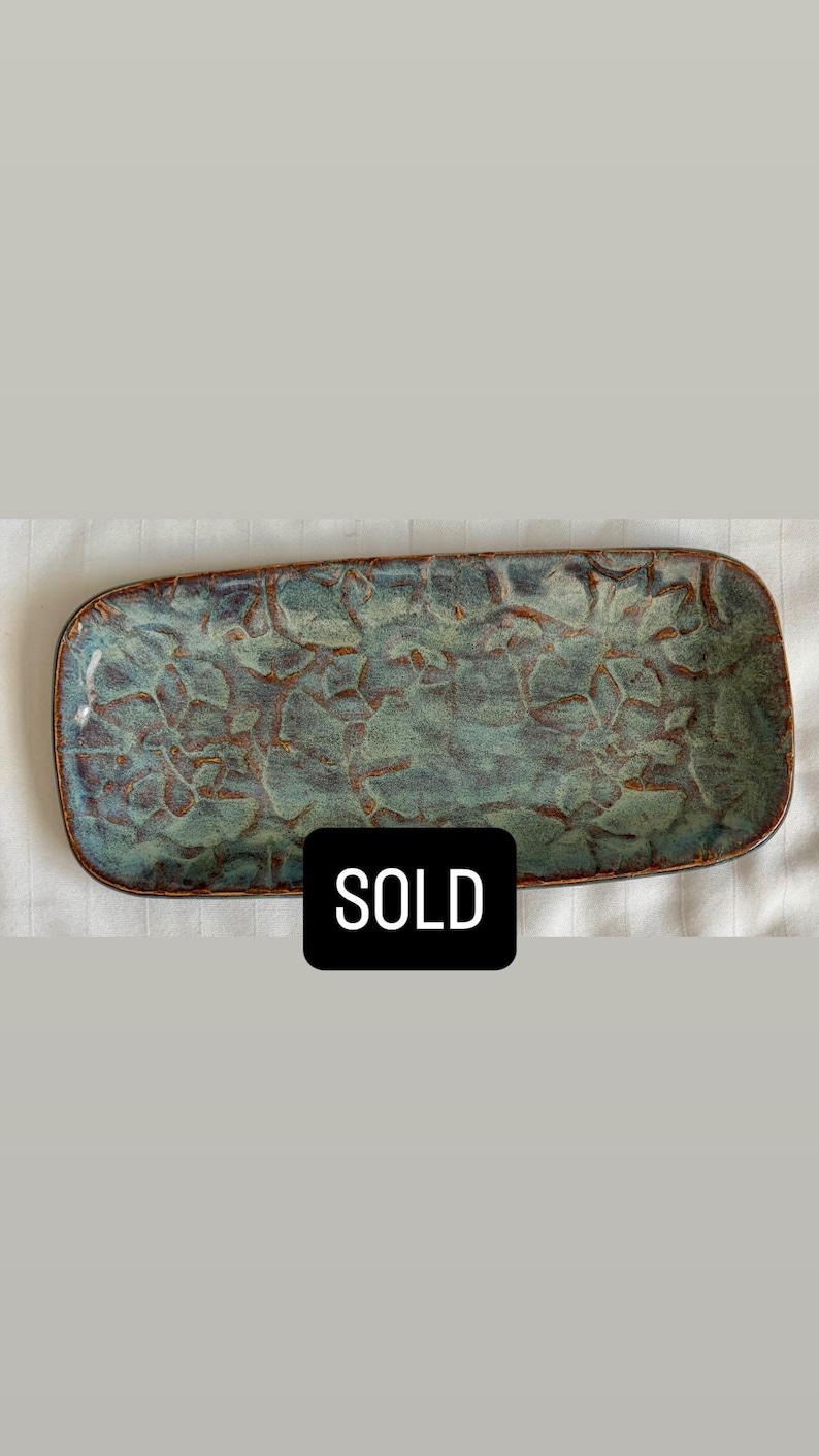 May include: A rectangular ceramic tray with rounded corners, featuring a mottled blue and green glaze with a textured, crackled surface pattern and a dark brown rim. A black rectangular label with the white text SOLD is centered on the tray.