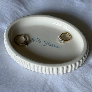 Peut inclure: Un vide-poche ovale en céramique blanche avec un bord extérieur cannelé. Le centre du plat présente le texte "The Sierras" en écriture bleue. Deux bagues en or, l'une avec une pierre sombre et l'autre avec une pierre claire, reposent à l'intérieur.