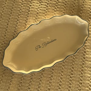Puede incluir: Una fuente de servir ovalada de cerámica color crema con un borde festoneado y un reborde gris oscuro. El centro del plato presenta el texto en cursiva negra "The Richardsons" impreso en medio de la superficie interior lisa y brillante.