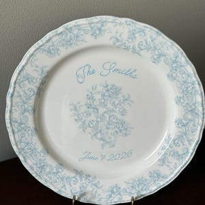 May include: A white ceramic plate with a scalloped edge, featuring a light blue floral pattern around the rim and centre. The text "The Smiths" is printed in blue script above the floral design, with "June 7 2026" printed in blue below it.