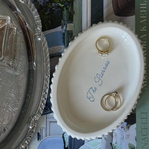 Puede incluir: Un plato ovalado de cerámica blanca con borde de cuentas y el texto azul en cursiva The Sierras impreso en el centro. Tres anillos de oro descansan dentro del plato, colocado junto a una bandeja de plata con un borde ornamentado en relieve.