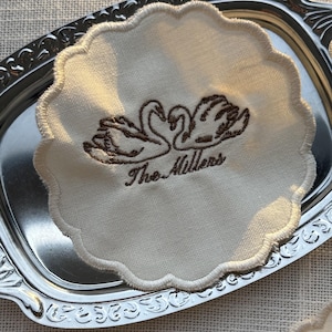 May include: A round, off-white fabric coaster with a scalloped edge, featuring brown embroidery of two swans facing each other and the text "The Millers" underneath. The coaster is placed on a small, rectangular, silver-coloured metal tray.