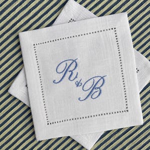May include: Two square white linen napkins featuring a decorative hemstitch border and a central blue embroidered monogram of the letters R and B connected by an ampersand. The fabric has a visible weave texture and clean, straight edges.