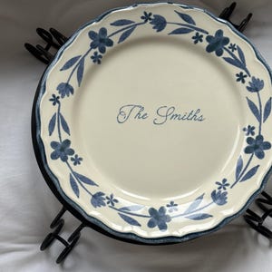May include: A round, cream-coloured ceramic plate with a scalloped edge and a blue floral vine pattern around the rim. The centre of the plate features the text "The Smiths" in a blue cursive script. The plate rests on a black metal stand.